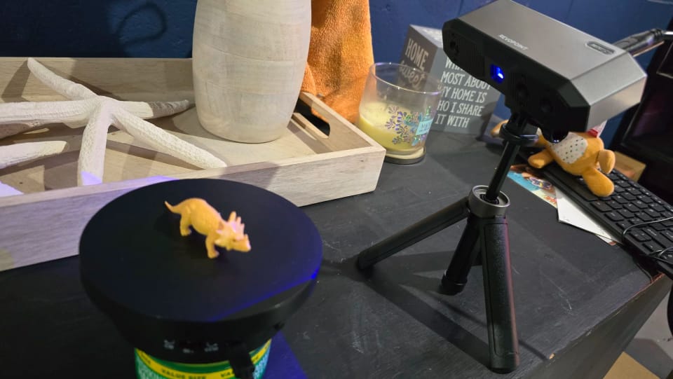 A 3D scanner on a tripod scanning a small figure on a turntable